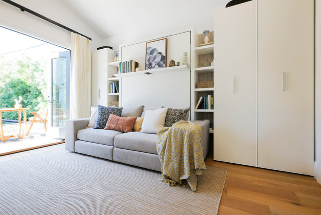 Modern living room with Resource Furniture Tango Sofa wall bed, light gray sofa with colorful pillows, built-in shelving and storage, and large glass doors opening to outdoor space.