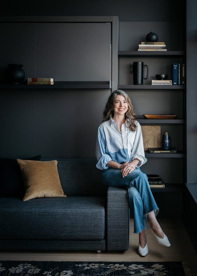 Seattle interior designer Michelle Dirkse seated on Oslo Murphy bed sofa in modern condo den with custom shelving and color-drenched walls.