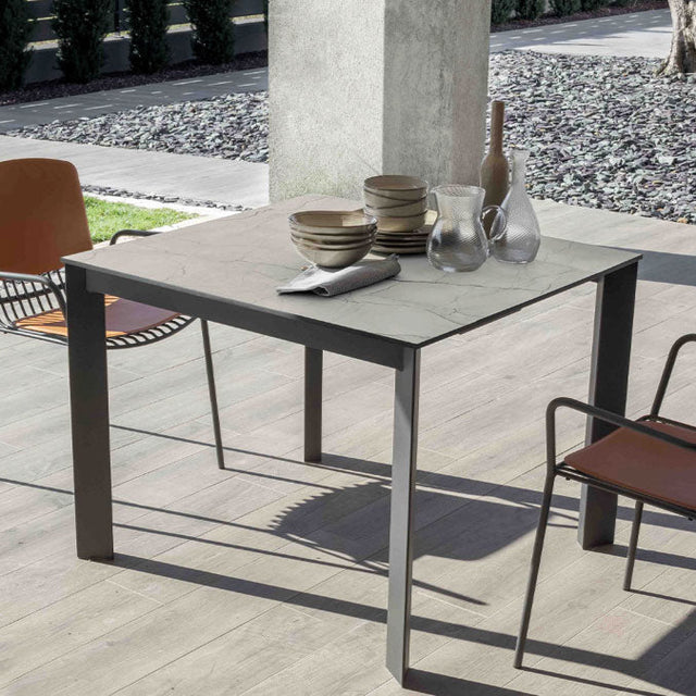 compact version of the Goliath Outdoor table with two orange and black chairs. The table is styled for casual dining with neutral-colored plates and glass pitchers.