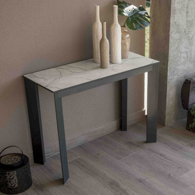 Another view of the Goliath Outdoor table in its console configuration. It is positioned on a patio against a textured wall, styled with decorative items, and paired with a black wire chair nearby.