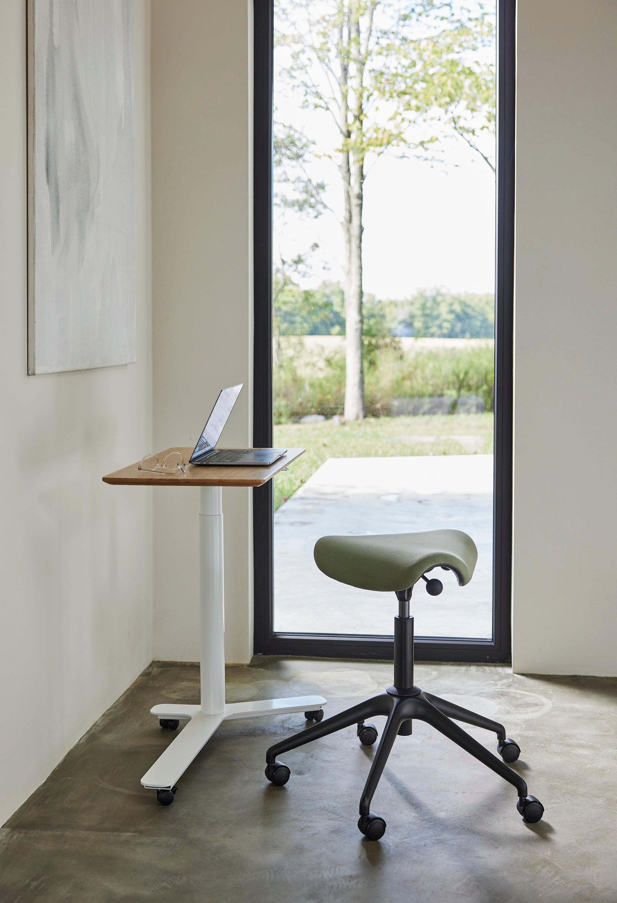 Float Mini height-adjustable desk with a green Pony saddle stool in front of a large window, ideal for light-filled workspaces.