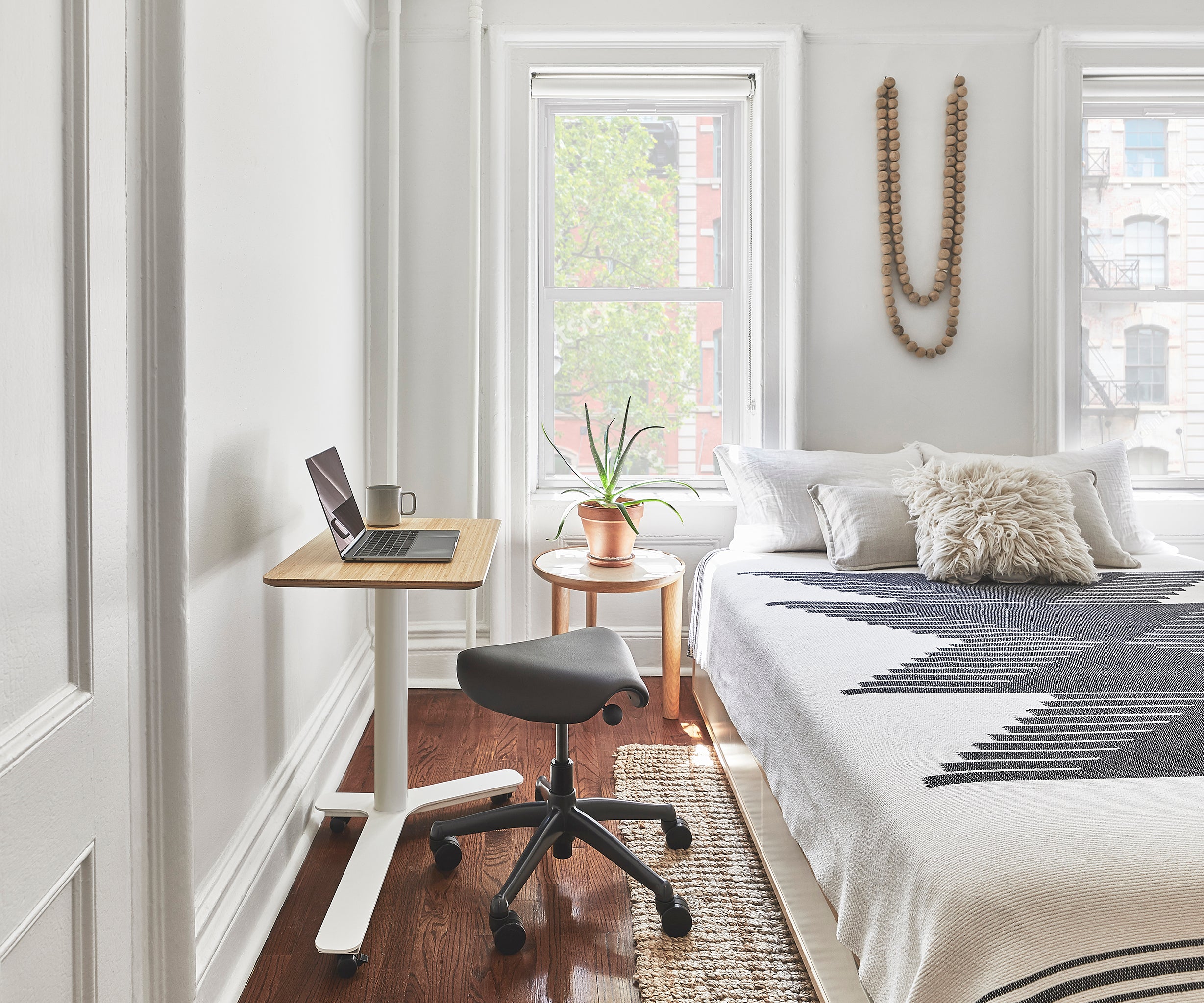 Compact Float Mini desk with bamboo top in a minimalist bedroom setting, paired with a black saddle stool and natural light from dual windows.