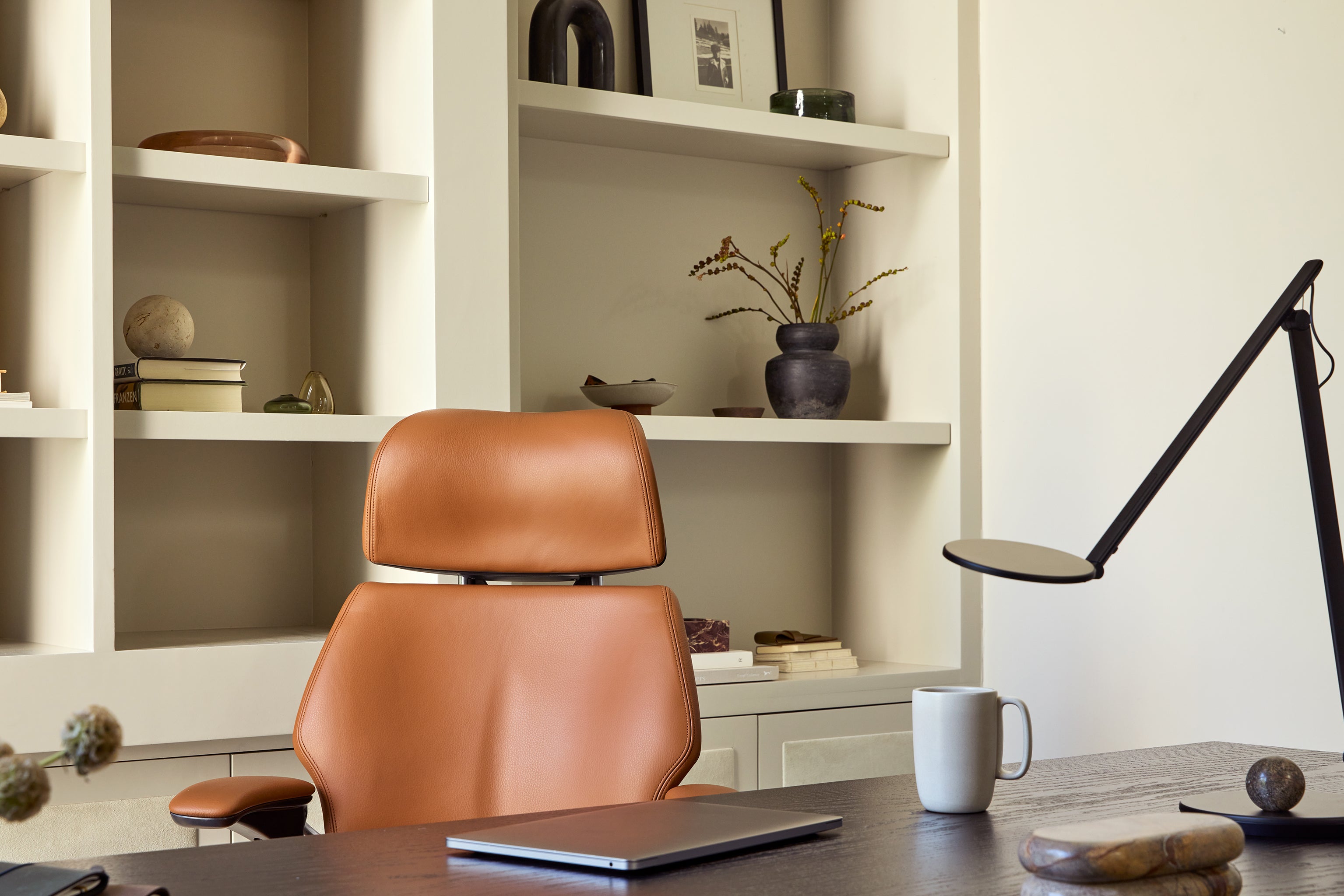 Close-up of tan leather Freedom Headrest chair in front of a modern desk, with shelving and task lighting in the background.