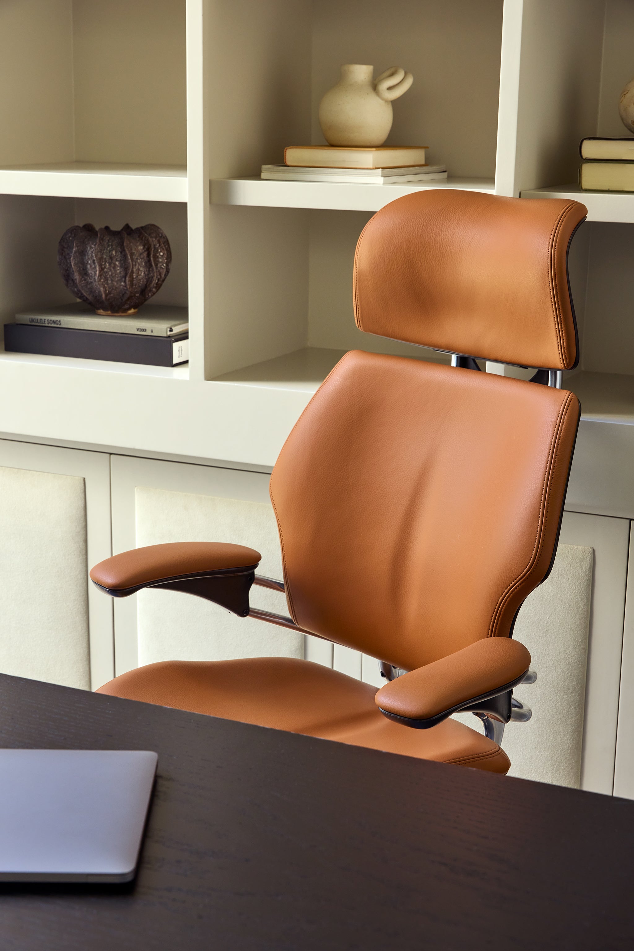 Tan leather Freedom Headrest ergonomic chair positioned at a desk with neutral shelving and sculptural decor in the background.