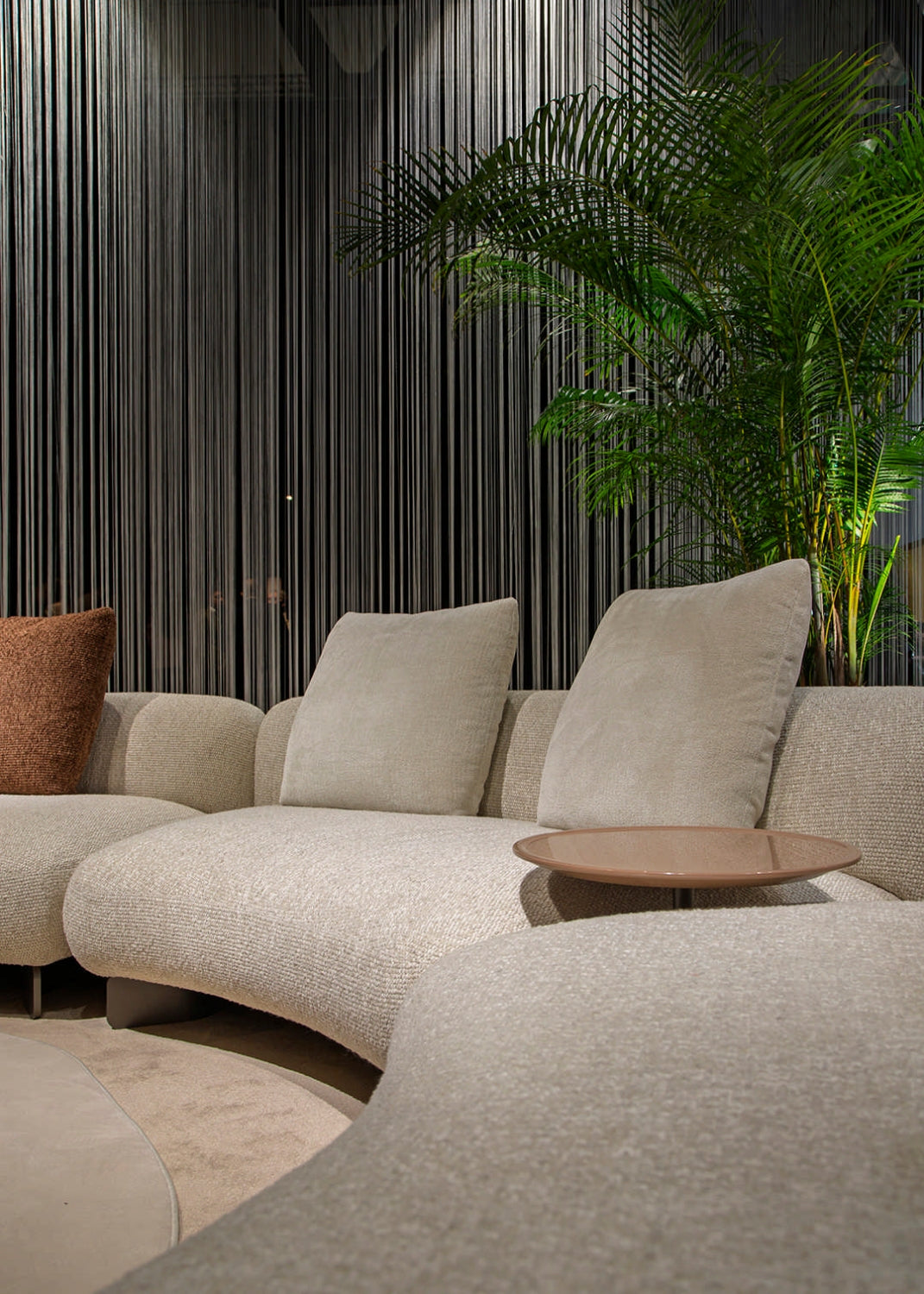 Detail view of the Kintai modular sofa showing soft curves, neutral fabric, and accent pillows beside greenery in a modern living room.