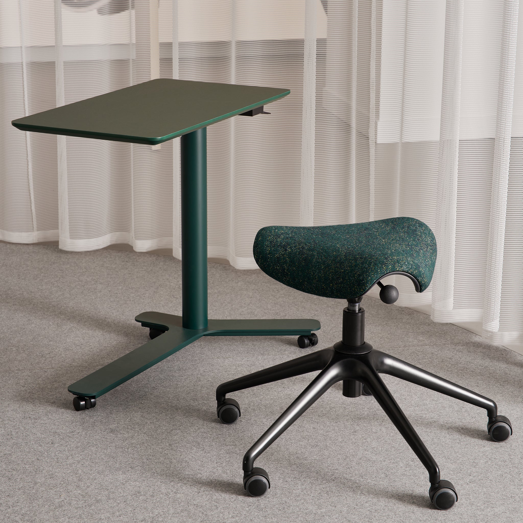 Ergonomic Pony saddle stool paired with a height-adjustable green side table, styled in a minimalist office with light curtains and grey carpet.