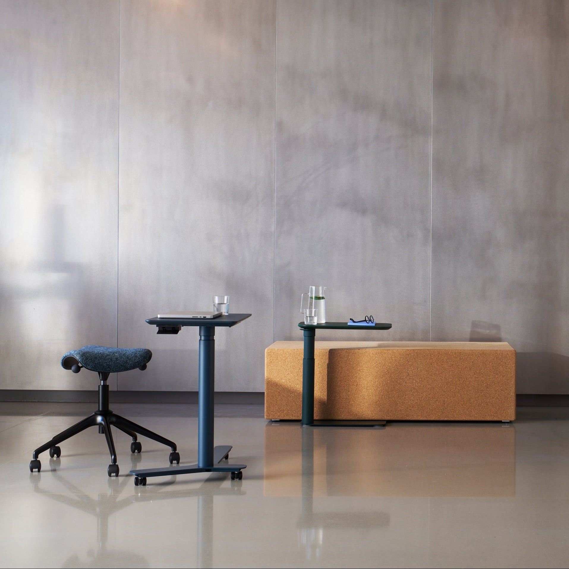 Modern lounge workspace featuring the Pony saddle stool and mobile height-adjustable side tables set against cork benches and grey walls in an open-plan office.