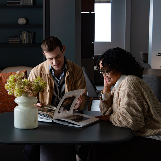 Two people sit at a Dynamic table in the Resource Furniture LA showroom, browsing a design book. The modern space features a wall bed with bookshelf, a cozy sofa with textured pillows, and soft, natural lighting.