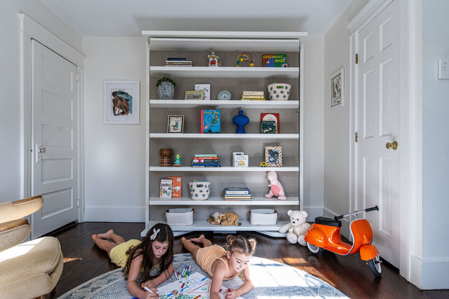 LGM Murphy wall bed (Resource Furniture) folded as white shelving in a kids' room, two children drawing on the rug and an orange ride-on scooter beside the unit — space-saving, multifunctional furniture for small bedrooms.