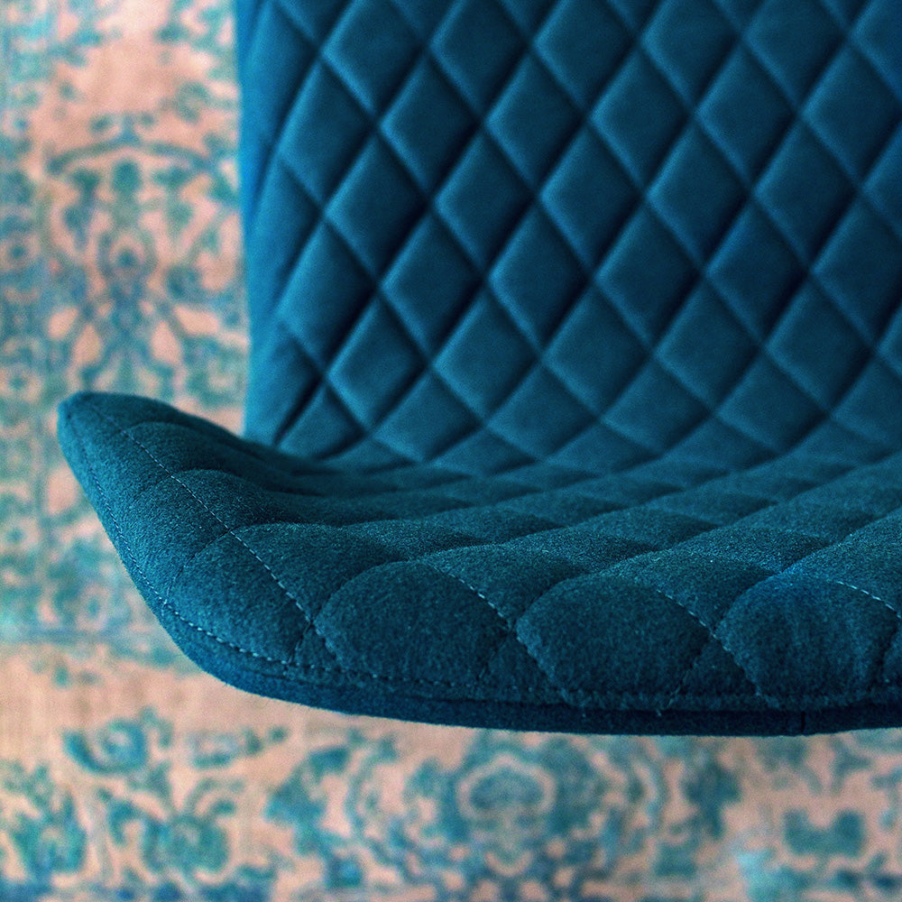 Detail of Ava dining chair’s diamond-quilted teal upholstery, showing stitching and curved seat edge against a patterned rug.