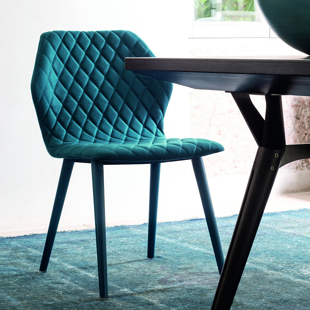 Ava dining chair in teal quilted upholstery with matching wood legs, shown beside a dark-stained dining table in a sunlit interior.