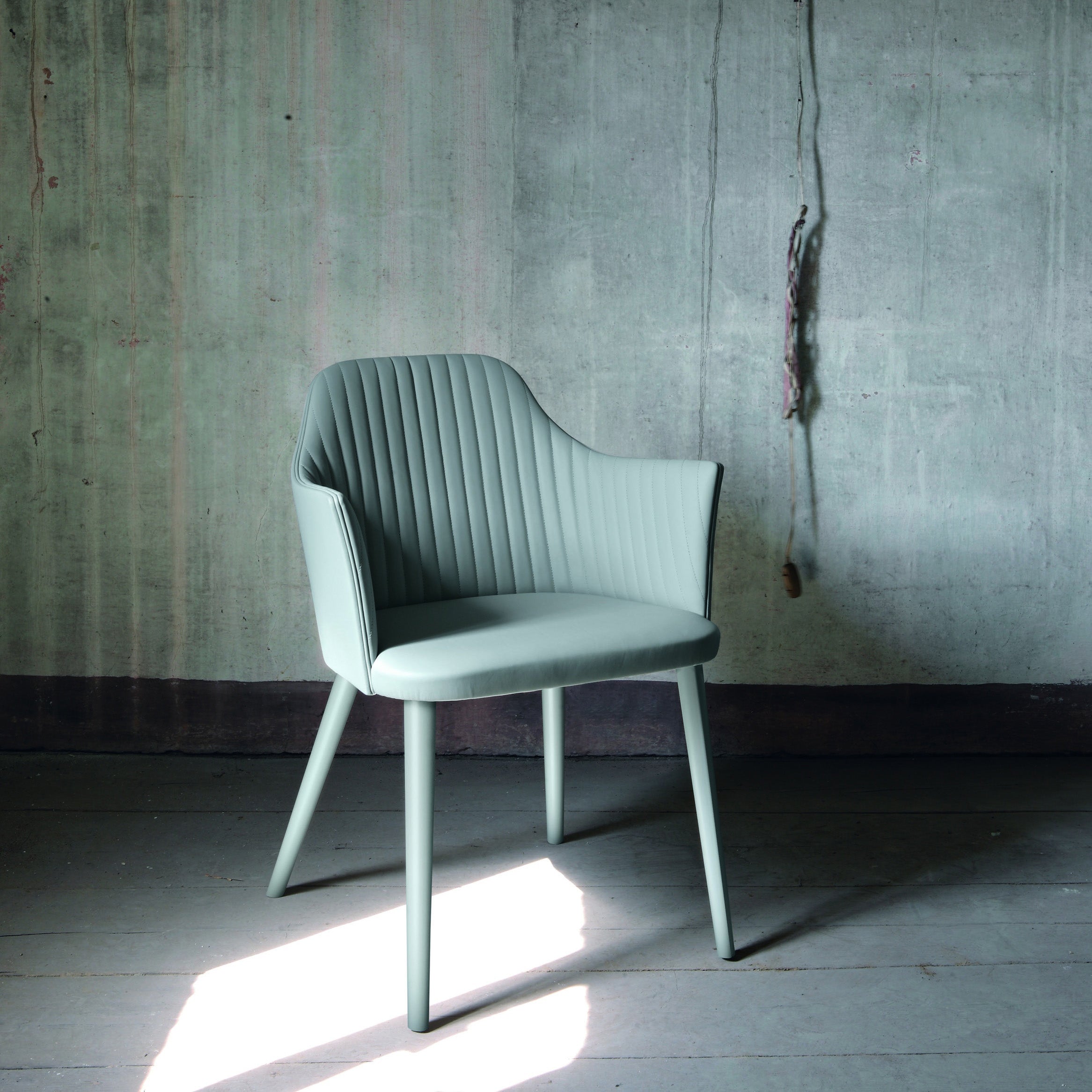 Break Dining Chair in light green upholstery with vertical quilting, armchair version shown against a raw concrete backdrop.