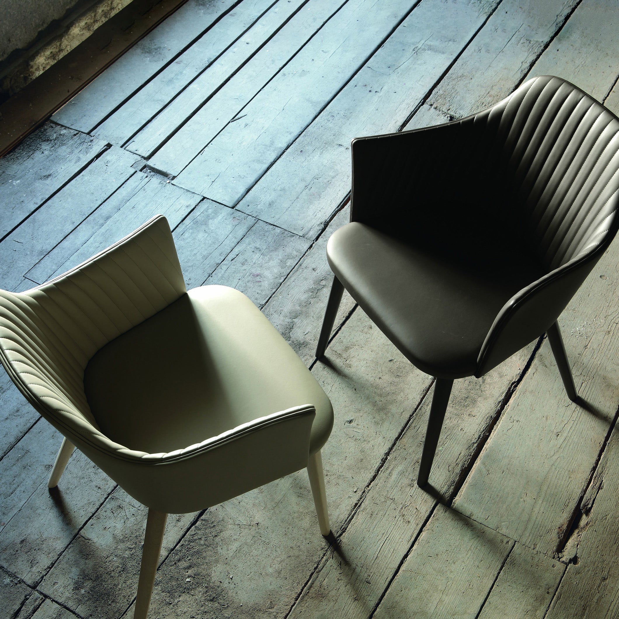 Overhead view of two Break Dining Chairs, one in olive and one in black upholstery, showcasing quilted backs and wooden legs on rustic plank flooring.