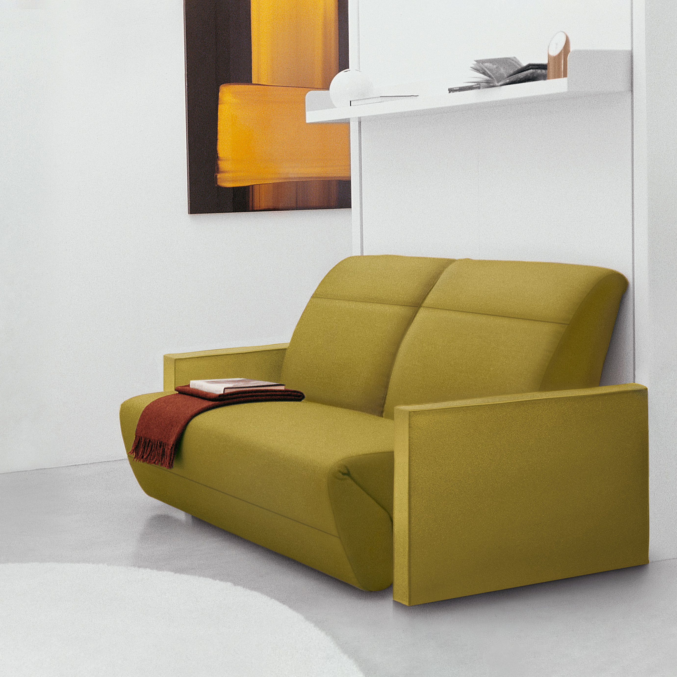 Ito wall bed closed, configured as a reclining sofa in green fabric with a red throw, situated in a contemporary white room with floating shelf above.