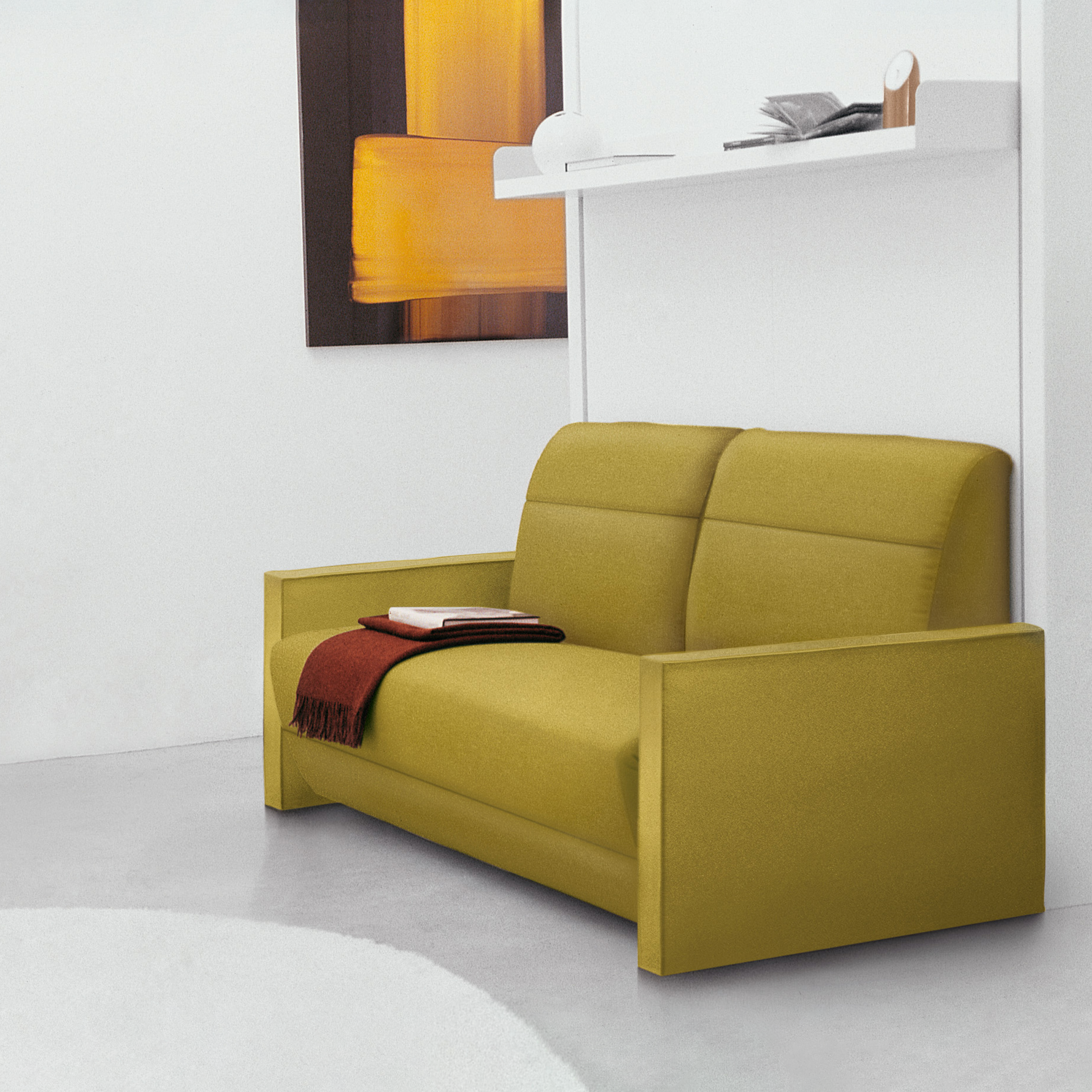 Ito wall bed closed, configured as a reclining sofa in green fabric with a red throw, situated in a contemporary white room with floating shelf above.