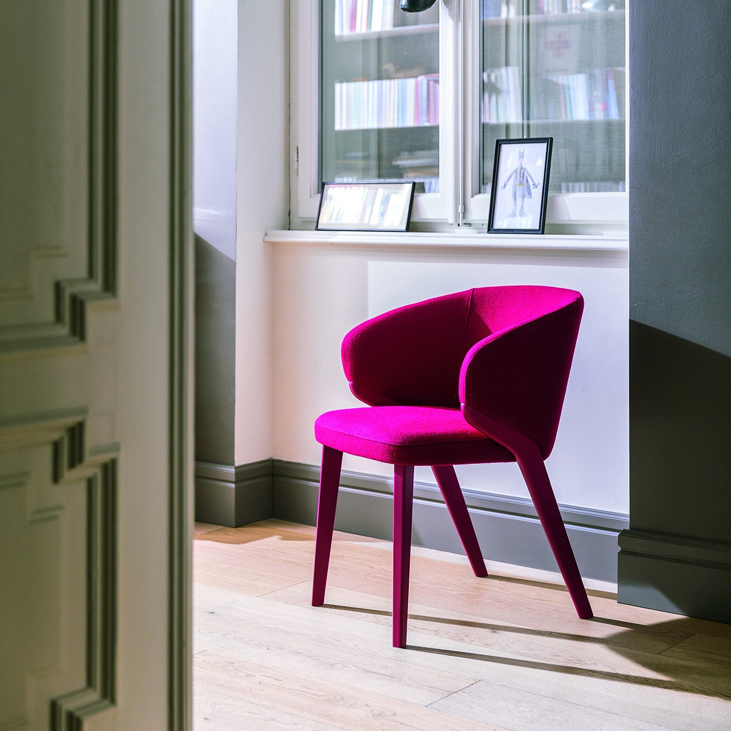 Nora dining chair in magenta upholstery with wraparound backrest, shown in a residential interior with wood flooring and natural light.