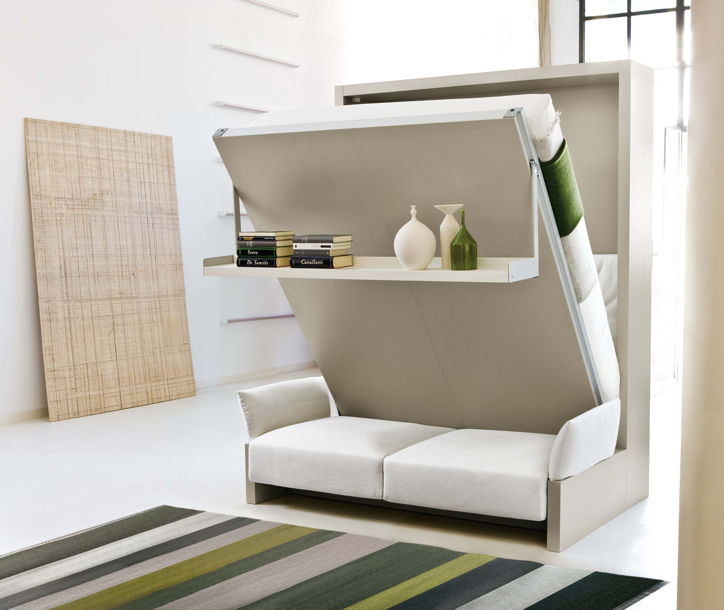 Nuovoliola wall bed in mid-transition from sofa to queen-size bed, featuring a pivoting shelf that stays level while opening, with white upholstery and green-accented bedding—ideal for multifunctional furniture in small spaces.