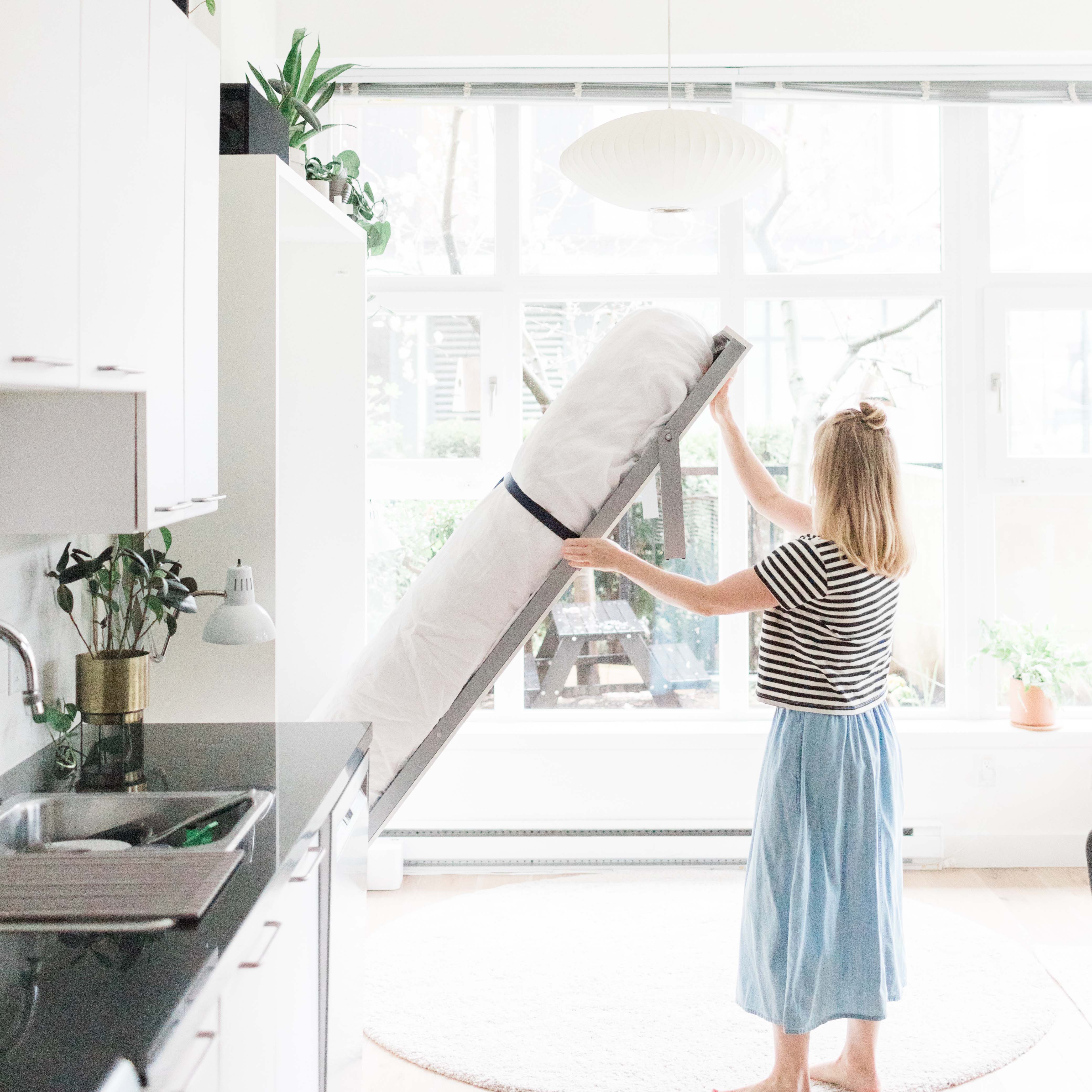 Woman unfolding a Penelope queen Murphy bed in a bright, minimalist kitchen with large windows.
