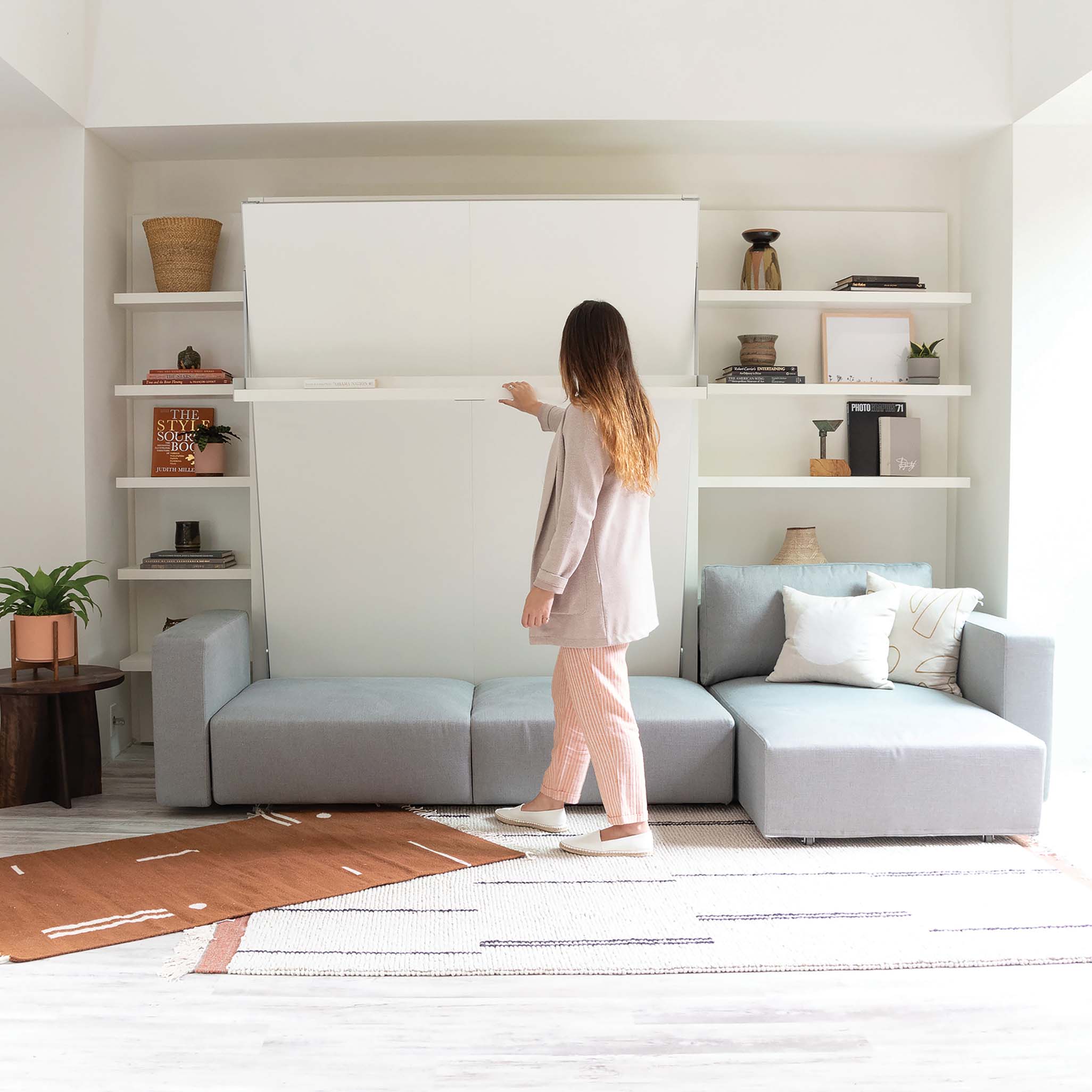 Woman opening the Swing Chaise wall bed in a modern living room with built-in shelving and a modular sofa.