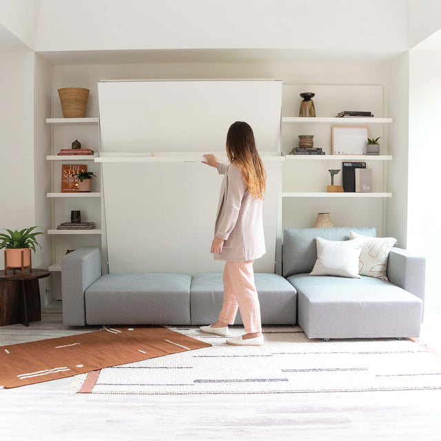 Woman opening the Swing Chaise wall bed in a modern living room with built-in shelving and a modular sofa.