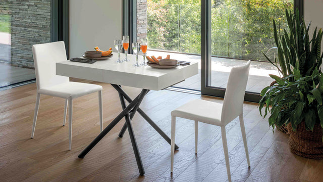 Brooklyn Mini table extended to dining height, set for two with white chairs, against a backdrop of greenery and modern decor.