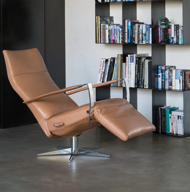 Cascade modern recliner in caramel leather with a sleek chrome base, positioned in a contemporary living room with a bookshelf in the background.