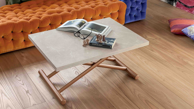 Doppio Long transforming table in its closed configuration, styled with a beige ceramic top and bronze base, in a vibrant living room with orange tufted seating.