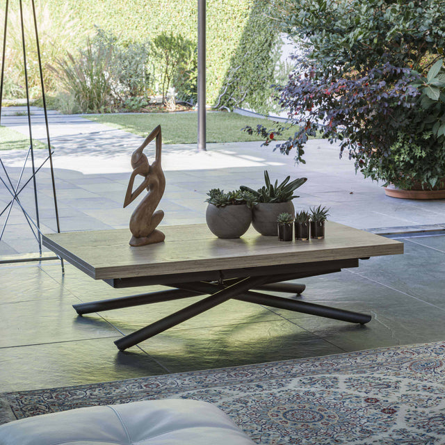 The Rotasu transforming table in its coffee table configuration, set against an outdoor patio backdrop featuring lush greenery and sculptural decor.