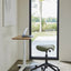 Float Mini height-adjustable desk with a green Pony saddle stool in front of a large window, ideal for light-filled workspaces.