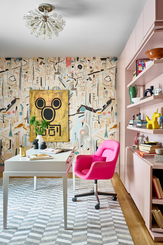 LGM wall bed closed as a sleek millennial-pink media wall, styled with open shelving, art books, and a white desk with a bright pink swivel chair in front of a bold abstract wallpaper.