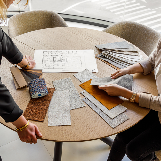 Two people compare upholstery fabric and leather swatches over a round oak table with a printed floor plan at the Resource Furniture NYC showroom. 