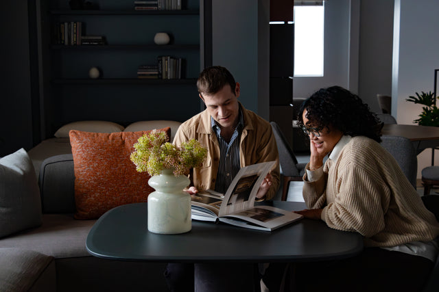 Two people sit at a Dynamic table in the Resource Furniture LA showroom, browsing a design book. The modern space features a wall bed with bookshelf, a cozy sofa with textured pillows, and soft, natural lighting.