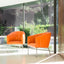 Two vibrant orange Rumba chairs with quilted leather upholstery and sleek chrome legs placed in a bright, modern space with large glass windows overlooking lush greenery.