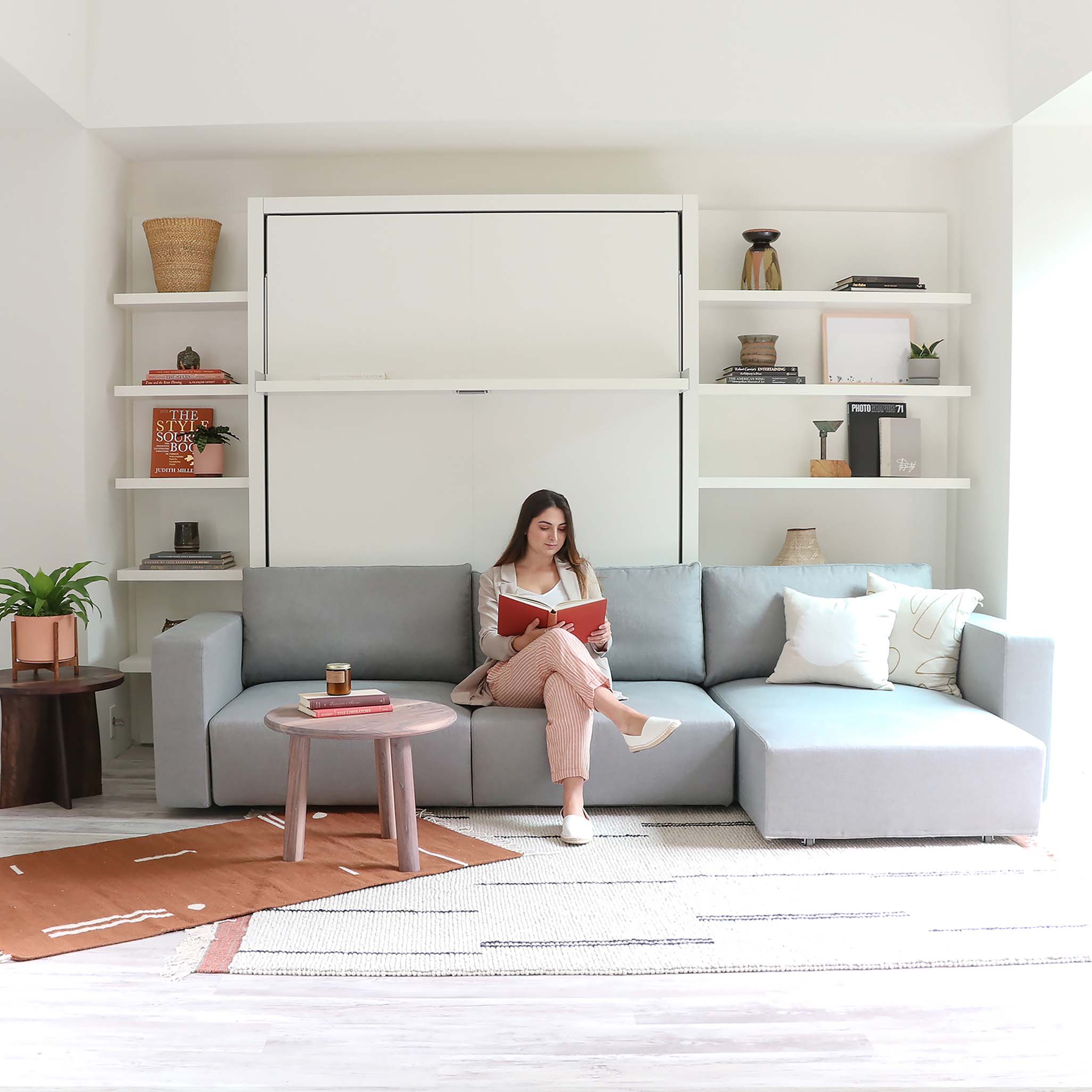 Woman reading on the Swing Chaise sofa in front of a closed Swing wall bed with built-in shelving.