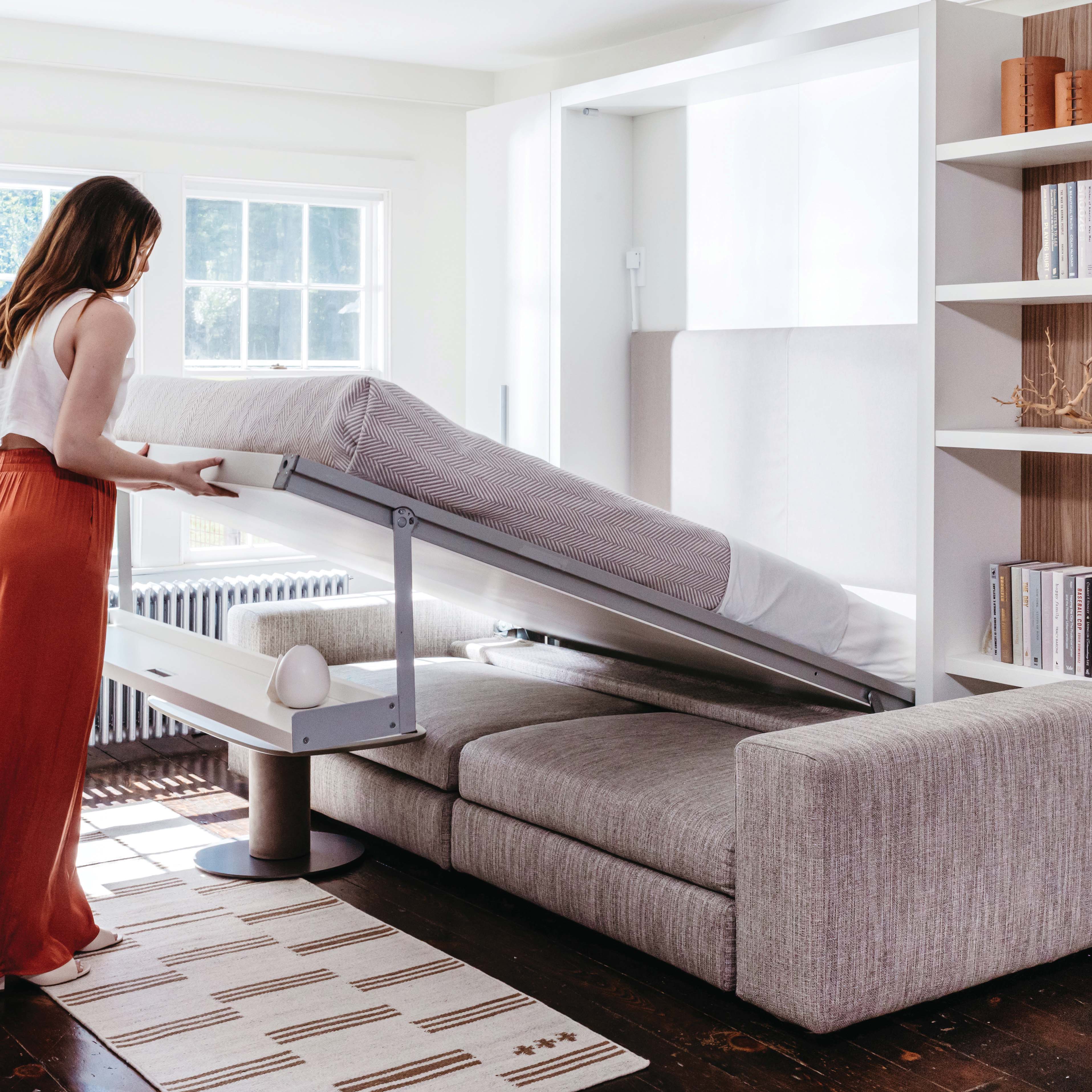 Woman lowering a queen-sized wall bed from the Tango Sofa system in a modern, multifunctional living room—ideal for small spaces and space-saving furniture solutions.